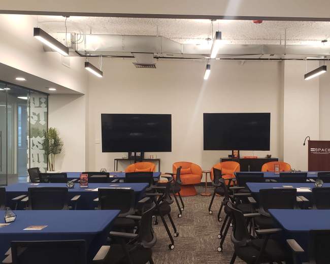 The image shows a modern conference room with blue tables arranged in rows, two large screens on the back wall, and orange chairs in the center.