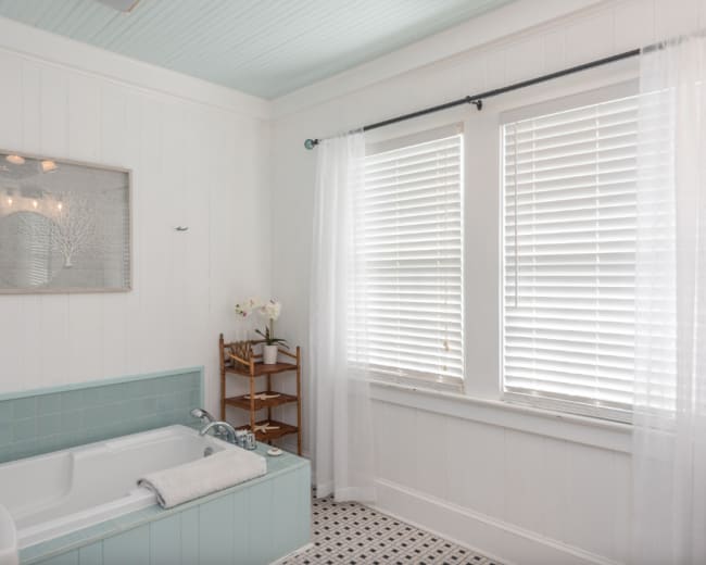 The bathroom features a light blue tiled tub, a wooden shelf with toiletries, and large windows with white blinds.