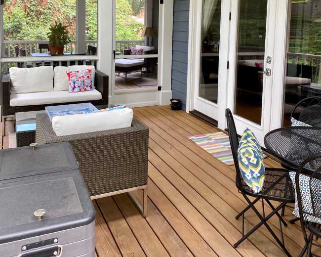 The image shows a screened porch with seating, a ceiling fan, and wooden flooring, surrounded by greenery.