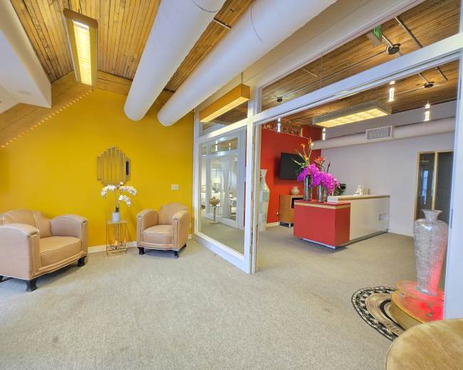 The image shows a modern reception area with two light-colored chairs, a vibrant yellow wall, and an adjoining workspace featuring a red counter and floral decorations.