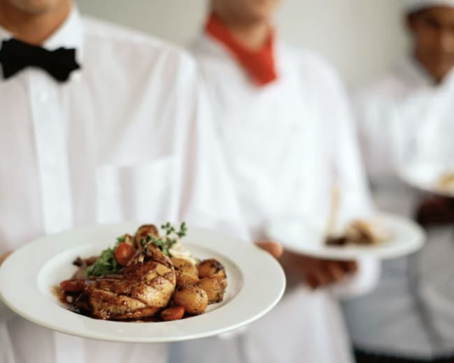 Three chefs in white uniforms hold plates of food, with one chef in the foreground presenting a dish featuring roasted chicken, potatoes, and garnishes.