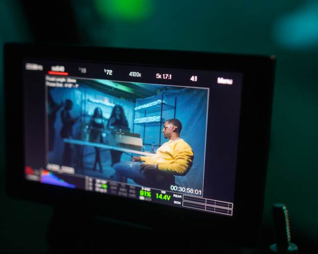 A monitor displaying a scene from a film set featuring a person sitting at a table while three others stand in the background.