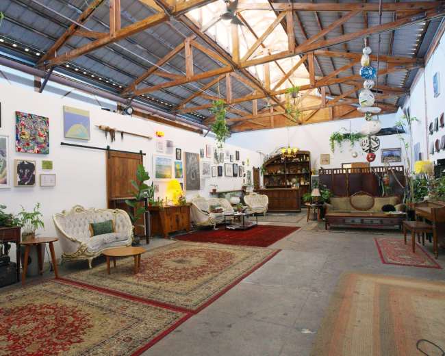 The image shows a spacious, well-lit room with wooden beams, decorated with various artworks on the walls and furnished with vintage seating and rugs.