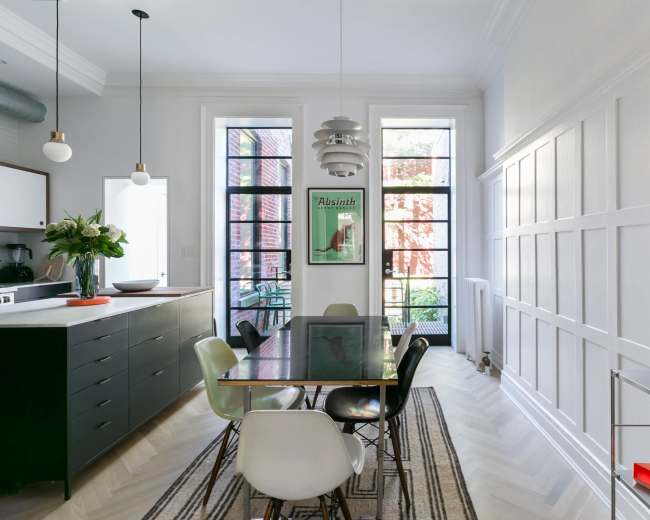 A modern dining area with a glass table surrounded by various colored chairs, large windows letting in natural light, and a minimalist kitchen in the background.