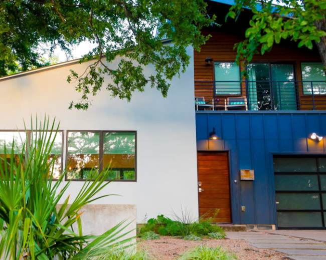 The image shows a modern two-story house with a mix of white and blue siding, surrounded by greenery and featuring large windows.