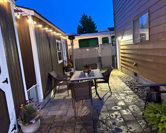 The image shows a well-lit outdoor patio area with a table and chairs, flanked by two buildings, and a pathway leading to a fenced section at the back.