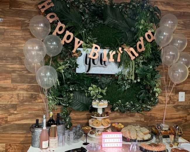The image depicts a birthday celebration setup featuring a table with various snacks and drinks, a "Happy Birthday" banner, and balloons against a green foliage backdrop.
