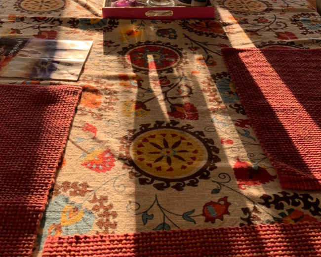 A dining table set with a patterned tablecloth, a pitcher, glasses, and various items, illuminated by sunlight coming through the glass doors.