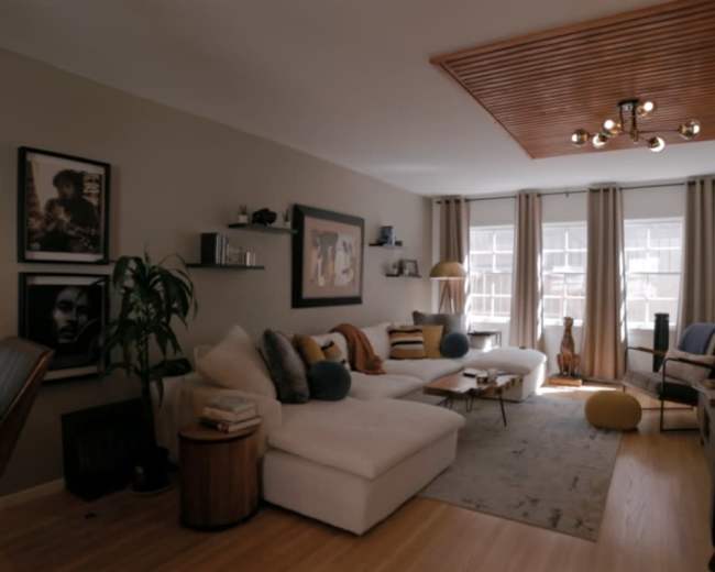 The image shows a modern living room with a white sectional sofa, a wooden ceiling feature, and large windows dressed with light curtains.