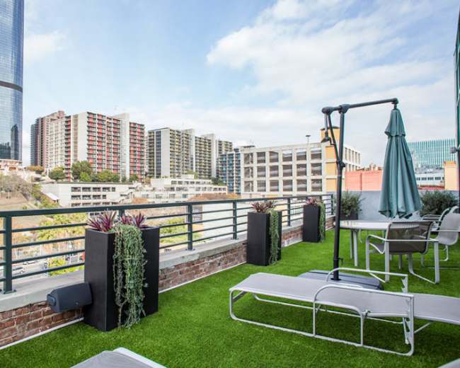 A rooftop terrace with lounge chairs and planters, overlooking a cityscape with tall buildings and greenery.