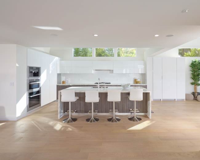 The image shows a modern kitchen with a central island, four white bar stools, and large windows allowing natural light to fill the space.