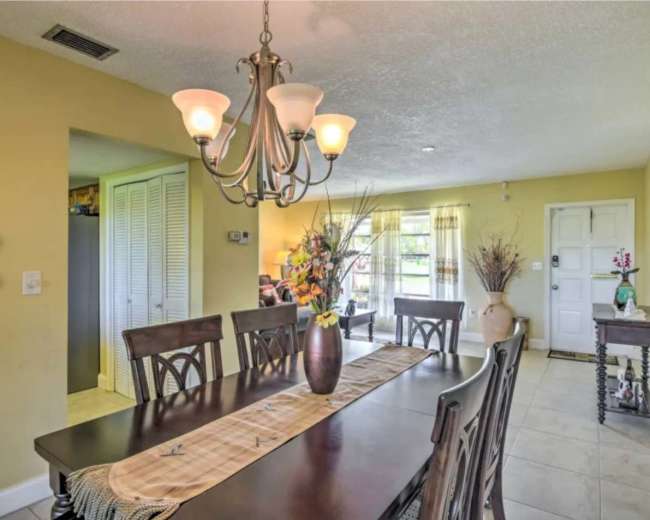 A dining area features a long wooden table with a vase of flowers and a chandelier above, beside a light-filled living space with large windows.