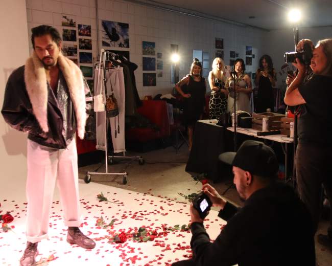 A male model in a fur-collared jacket stands on a carpet of rose petals while photographers capture the scene at a photoshoot attended by several spectators.