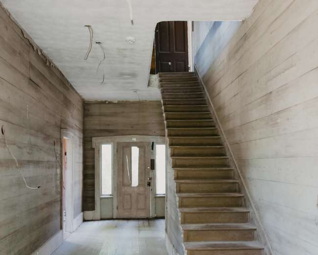 The image shows the interior of an unfinished house, featuring a staircase on the right and a doorway at the far end, with bare wooden walls and floor.