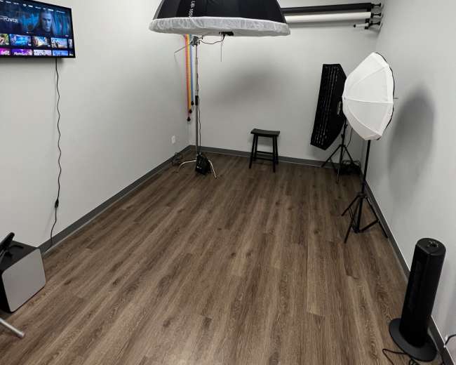 The image shows a small studio space with a light setup, a stool, and a television mounted on the wall.