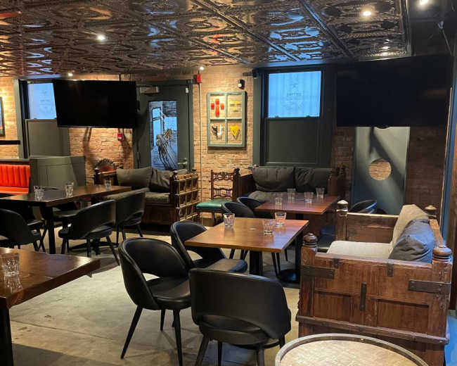 The image shows a dimly lit bar area with several tables and chairs arranged around a polished floor, featuring a decorative ceiling and exposed brick walls.