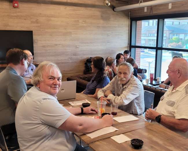 A group of people is seated at a long table in a casual dining setting, engaged in conversation and looking at menus.
