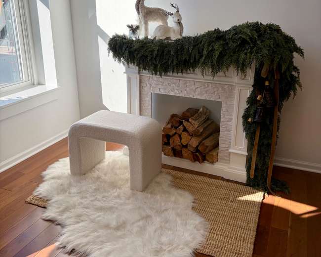 A white faux fur rug lies in front of a modern fireplace decorated with greenery and ornaments, accompanied by a minimalist stool and a stack of firewood.