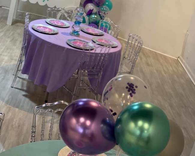 A decorated party space with tables set with plates and balloons, featuring a display cart in the background.