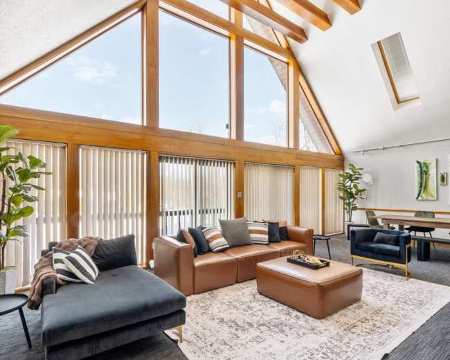 The image shows a spacious living area with large windows, wooden beams, and modern furniture including a sectional sofa and coffee table.