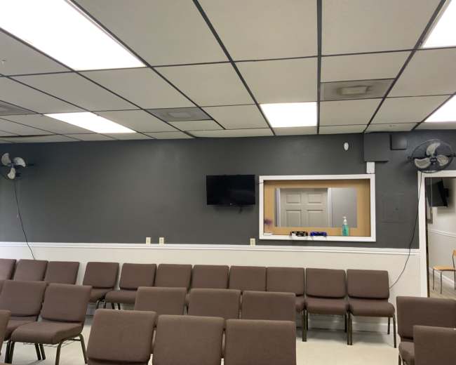 The image shows a seating area in a room with gray walls, a television mounted on the wall, and a small window to another room.