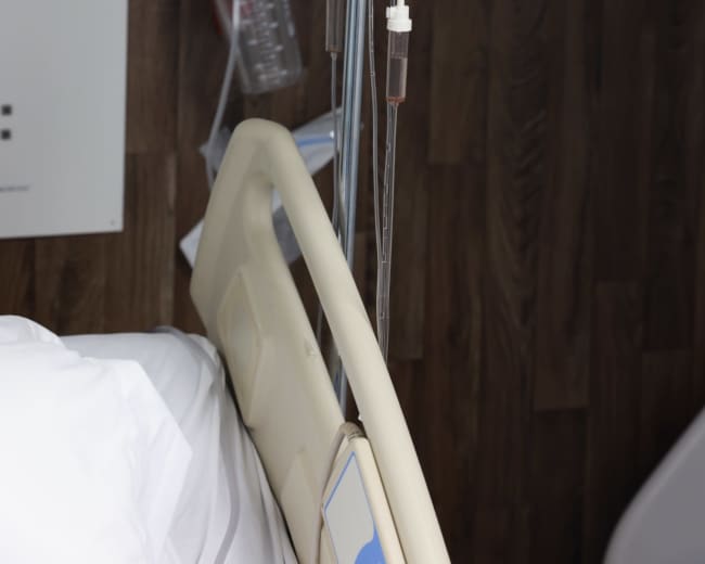 An IV stand with two bags of fluid is positioned next to a hospital bed, against a wood-paneled wall.