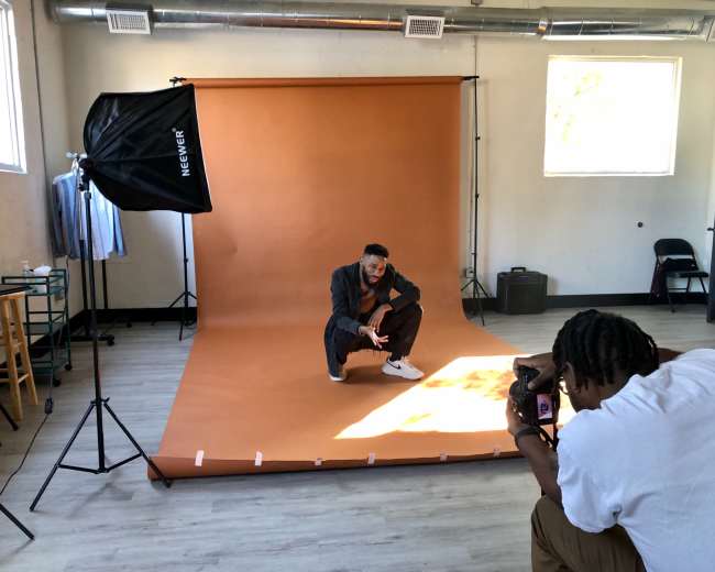 A photographer captures an image of a model squatting on a brown backdrop in a well-lit studio.