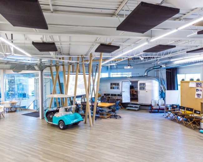 The image shows a spacious, modern office area featuring wooden pillars, a light blue golf cart, and a vintage trailer alongside several tables and chairs arranged for collaborative work.