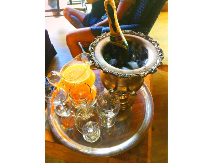 A silver ice bucket filled with ice holds a bottle of champagne, accompanied by various glassware and a pitcher of orange juice on a serving tray.