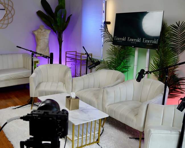 The image shows a modern studio setup with white chairs, a marble coffee table, a camera on a tripod, and colorful lighting accents.