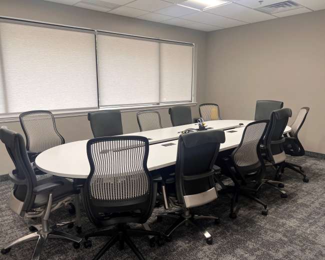 A large, oval conference table is surrounded by ten office chairs in a well-lit meeting room with window shades drawn.