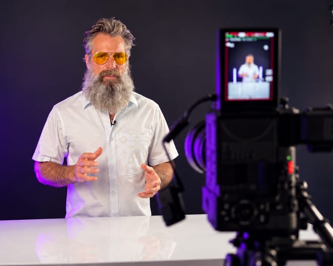 A man with a beard and yellow sunglasses gestures while standing behind a white table in front of a camera on a tripod.