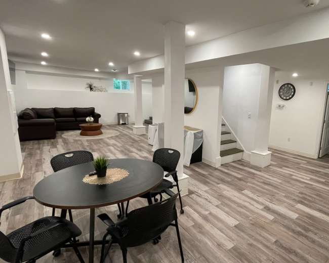 A modern, open-concept basement living space featuring a seating area, a dining table, and light-colored wooden flooring.