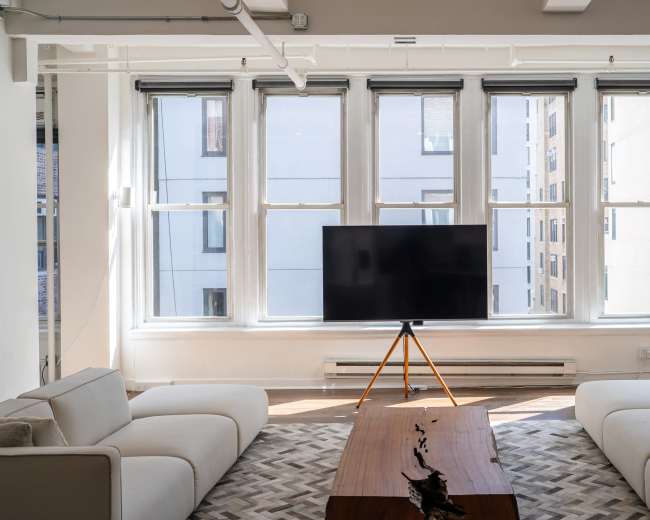 A modern living room features two white sofas facing a wooden coffee table, with a large television on a tripod stand positioned in front of windows that allow natural light to fill the space.