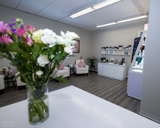 The image shows a modern aesthetic clinic interior with white furniture, a display area for skincare products, and a floral arrangement in a glass vase on a reception desk.