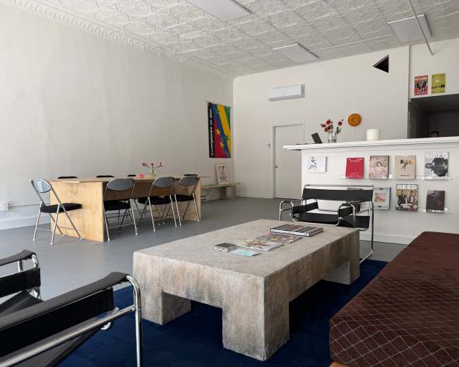 The image shows a minimalist interior of a room featuring a rectangular coffee table, a seating area with chairs, and a wall displaying magazines and posters.