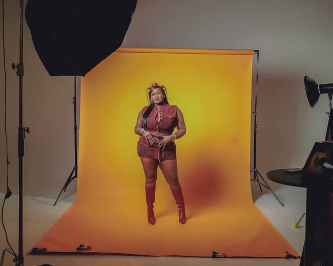 A model poses against a vibrant orange backdrop in a studio setting, illuminated by professional lighting equipment.