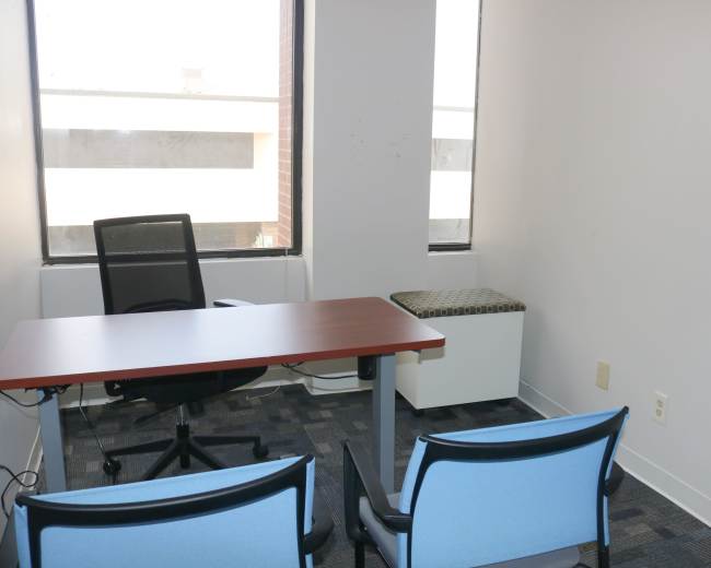 The image shows a small office space with a wooden desk, a black office chair, and two blue chairs facing the desk, along with a cushioned window seat.