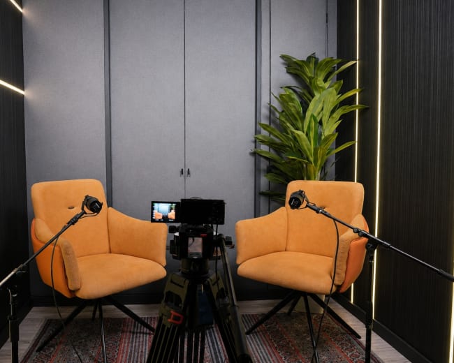 A video recording setup features two orange chairs facing a camera on a tripod, with a plant and acoustic panels in the background.