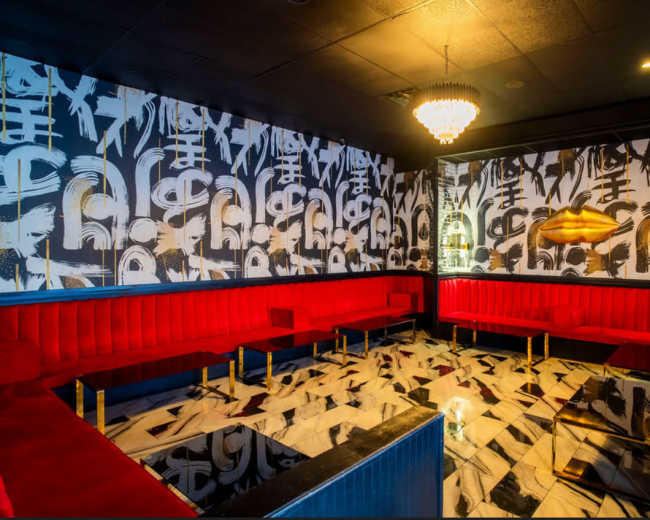 The image shows a chic lounge with red velvet seating, a marble floor, and a decorative chandelier.