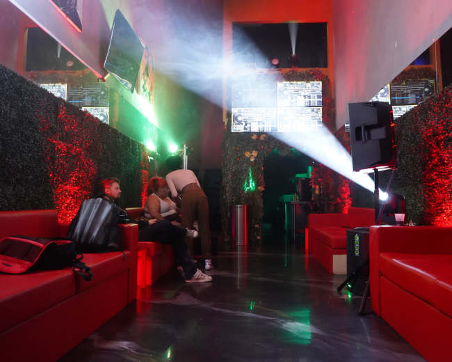 The image shows a dimly lit lounge area with red couches, greenery on the walls, and a DJ setup at the back, illuminated by colorful lights and projections.