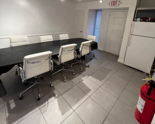 A long table with several white chairs is set up in a brightly lit room, featuring tile flooring and an exit door visible in the background.