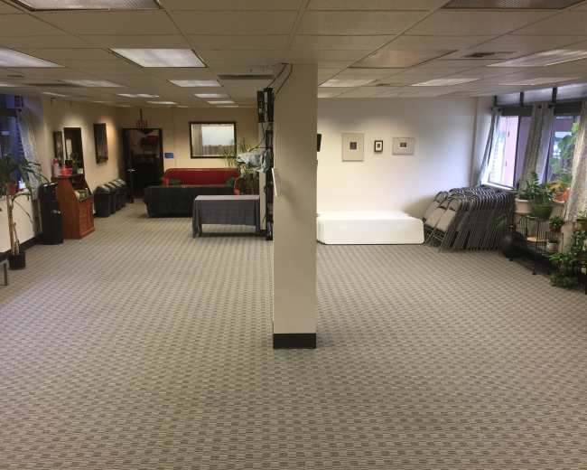 A spacious room with a divided layout, featuring seating areas and stacked folding chairs against the walls.