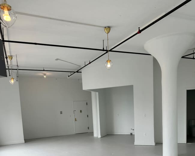 The image shows a spacious, empty room with gray flooring, white walls, and exposed lighting fixtures.