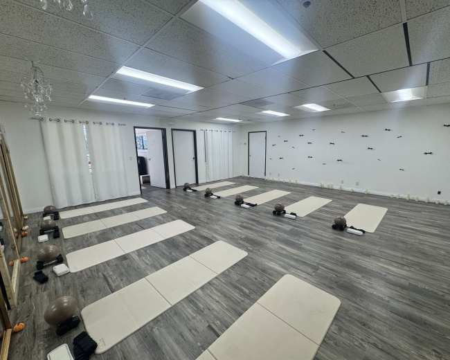 The image shows a spacious, uncluttered yoga studio featuring several exercise mats arranged in rows, with mirrors and soft lighting illuminating the area.