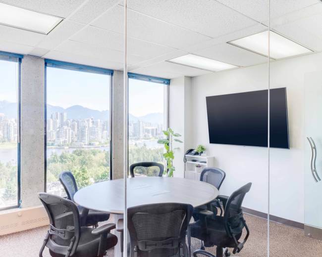 A modern meeting room features a circular table surrounded by chairs, with large windows offering a view of a city skyline and mountains in the background.