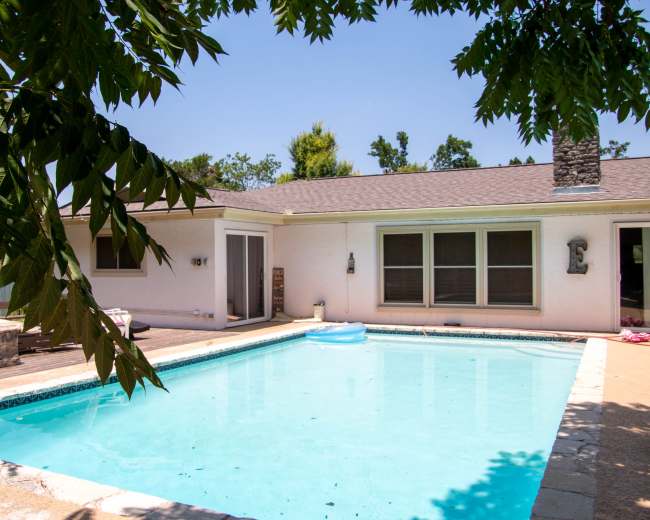 A clear swimming pool surrounded by a patio and a house with large windows on a sunny day.
