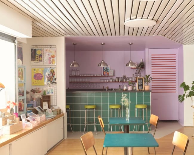 The image shows a modern café interior with a green tile counter, colorful walls, and wooden tables and chairs.