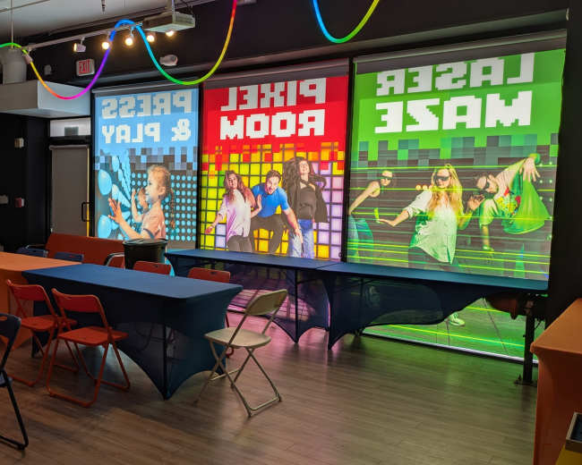 The image shows an indoor space set up for an event, featuring colorful tables and chairs, along with large, vibrant graphics on the windows.