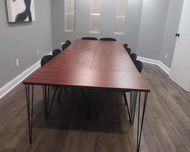 A long wooden table with metal legs is surrounded by black chairs in a well-lit, gray-walled meeting room.